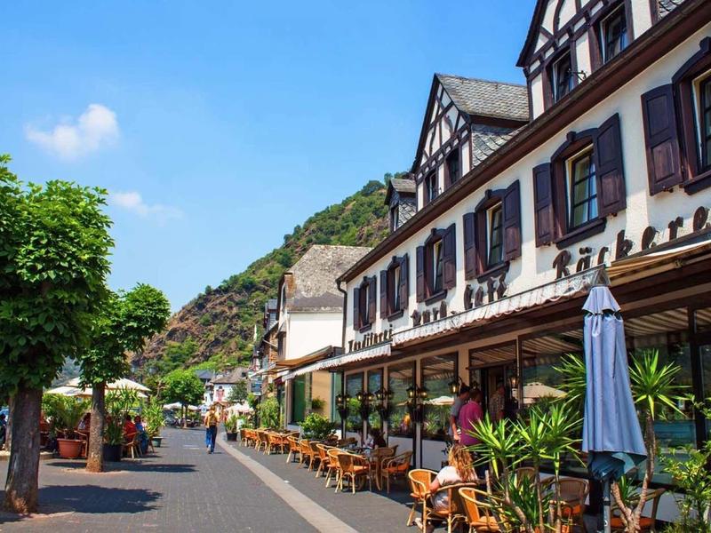 Straße mit Außengastronomie, alten Fachwerkhäusern, Bäumen und Hügel im Hintergrund bei blauem Himmel.