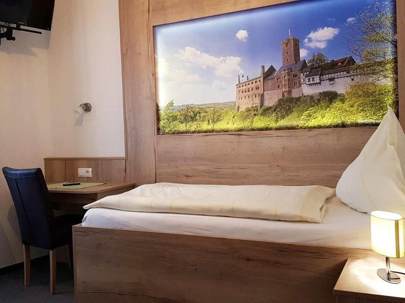 Kleines Zimmer mit Einzelbett, Schreibtisch, Stuhl, Fenster mit grüner Gardine und Wandbild einer Burg.