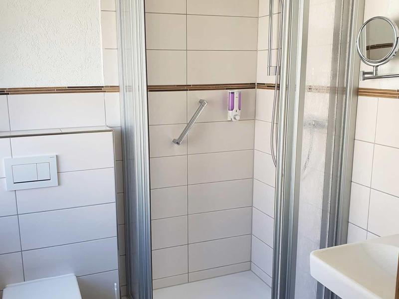 Kleine, helle Dusche mit Glasschiebetür, weißem WC, Waschbecken und braunem Boden im Badezimmer.