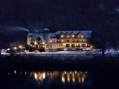 Beleuchtetes Hotelgebäude bei Nacht am Ufer eines ruhigen Gewässers im Winter.