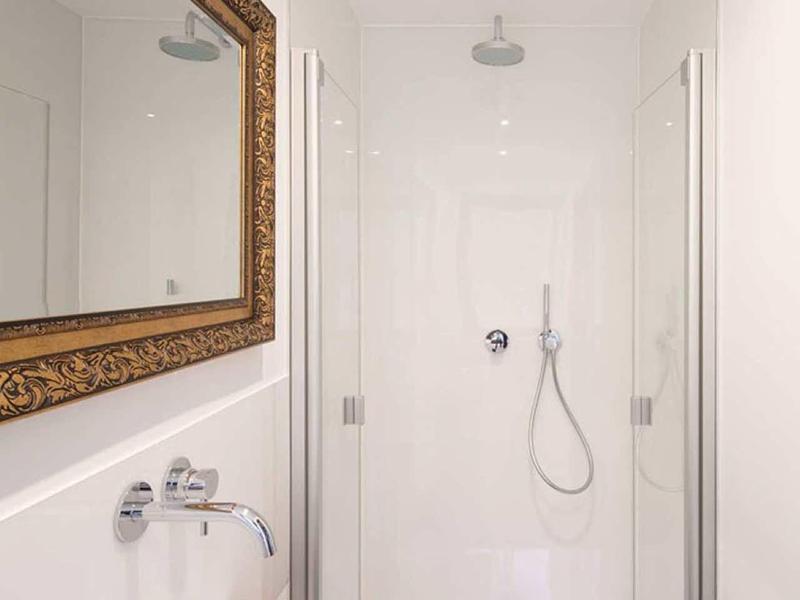 Schmale, helle Dusche mit weißer Wand, Regendusche, Handbrause und großem goldfarbenem Wandspiegel.