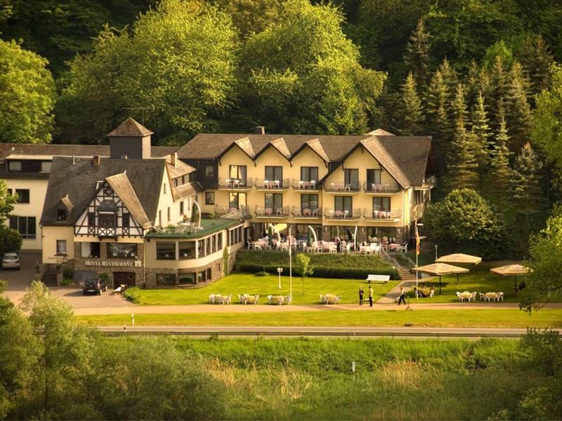 Großes Landhaus mit braunem Dach, Terrasse und Garten vor grünem Wald im Hintergrund.