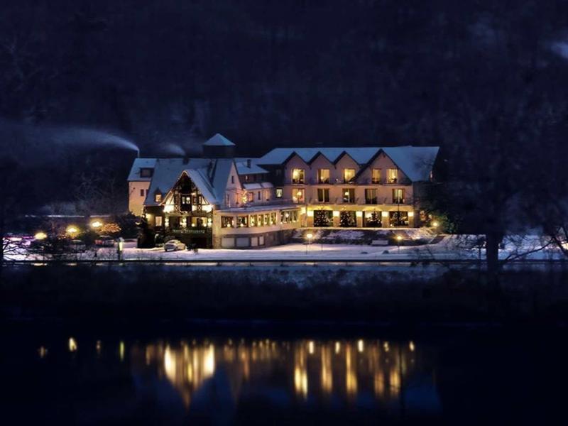 Ein beleuchtetes großes Haus am Ufer eines ruhigen Flusses bei Nacht, umgeben von dunklen Bergen.