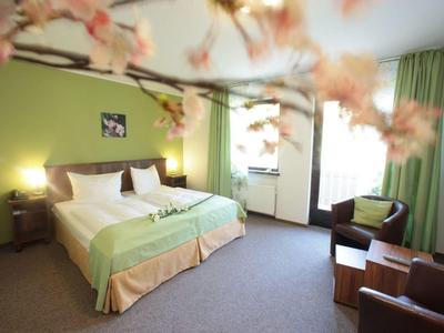 Hell grünes Schlafzimmer mit Doppelbett, Fenster, zwei Sesseln und blühendem Zweig im Vordergrund.