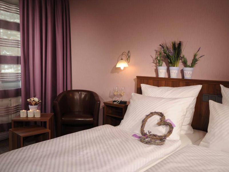 Gemütliches Schlafzimmer mit Doppelbett, weißen Kissen, braunem Sessel und violetten Vorhängen.