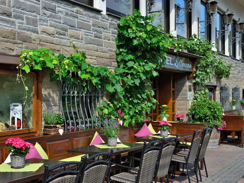 Außenterrasse eines Cafés mit gedeckten Tischen, grünen Pflanzen und Blumendekoration am Gebäude.