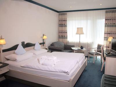 Hell eingerichtetes Hotelzimmer mit Doppelbett, Sessel, Fernseher und großem Fenster mit Gardinen.