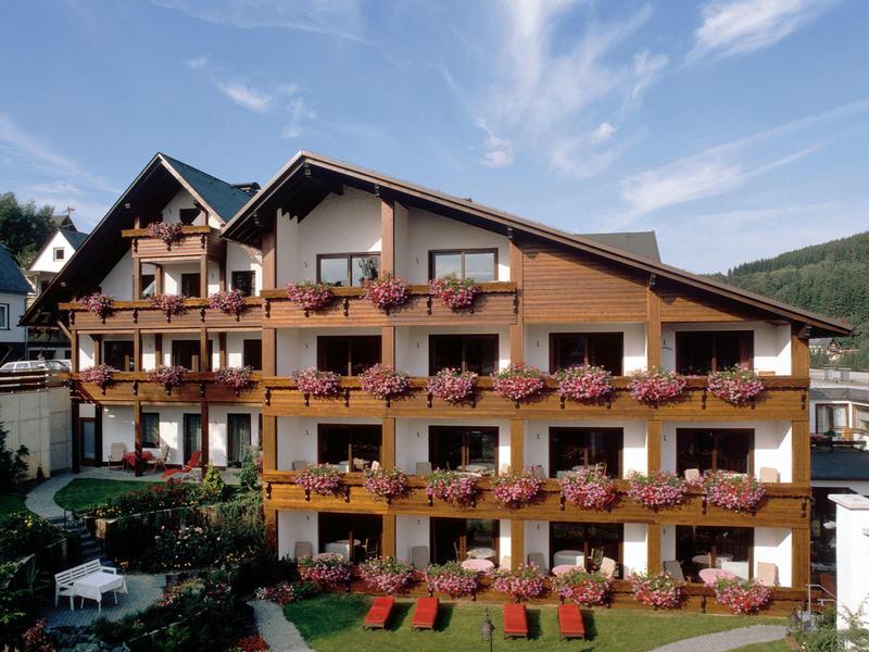 Großes alpenländisches Hotel mit Holzbalkonen und blühenden Blumen unter blauem Himmel.