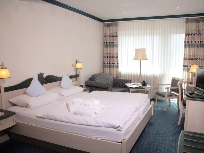 Hell eingerichtetes Hotelzimmer mit Doppelbett, Sessel, Fernseher und großem Fenster mit Gardinen.