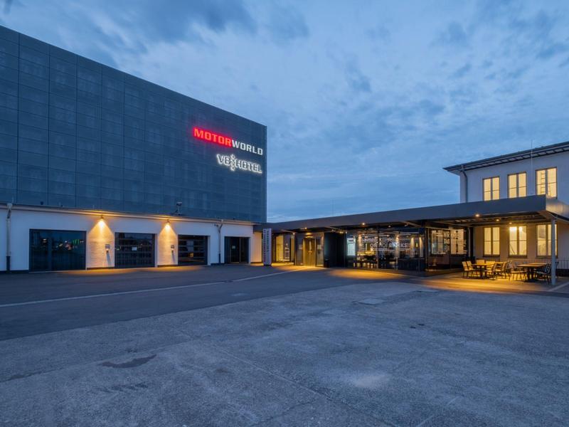 Modernes Gebäude mit beleuchtetem Eingang und großem Parkplatz bei Dämmerung und bewölktem Himmel.
