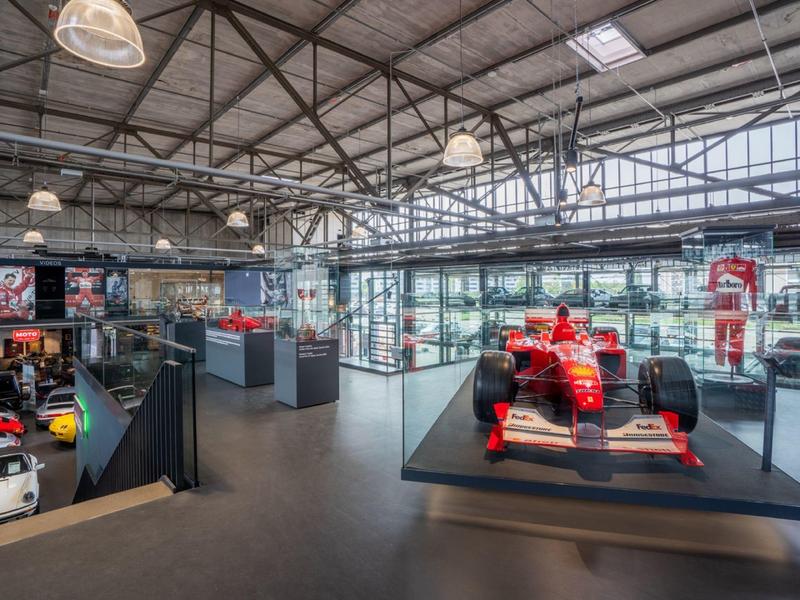 Moderne Ausstellungshalle mit rotem Formel-1-Rennwagen und weiteren Fahrzeugen hinter Glas.