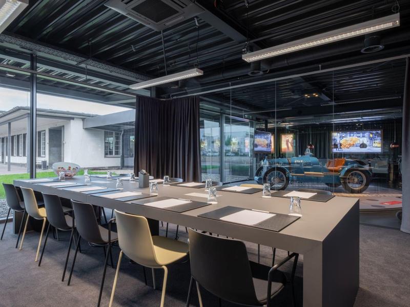 Moderner Besprechungsraum mit langem Tisch, bunten Stühlen und großen Fenstern mit Blick ins Freie.