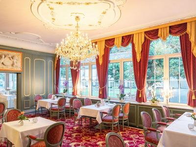 Eleganter Hotel-Speisesaal mit Kronleuchter, roten Sesseln und großen Fenstern mit roten Vorhängen.