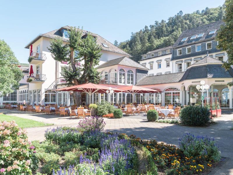 Hotelgebäude mit Terrasse, Sonnenschirmen und umliegendem Garten in sonniger Landschaft.