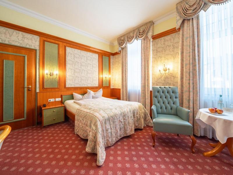 Elegantes Hotelzimmer mit Doppelbett, Sessel und Tisch vor großen Fenstern mit Vorhängen.