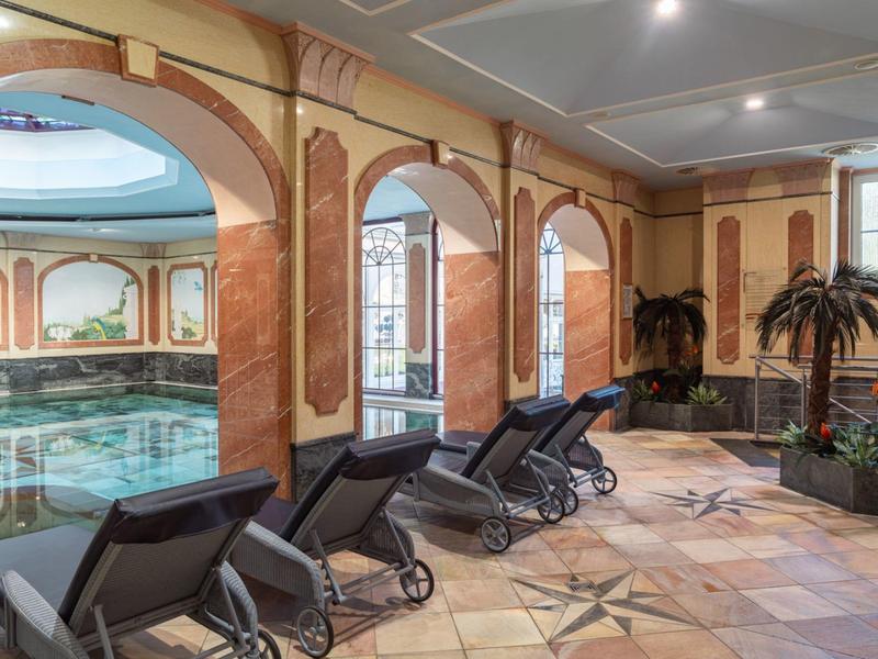 Innenbereich mit einem luxuriösen Hallenbad und Liegestühlen neben dekorativen Säulen und Fenstern.