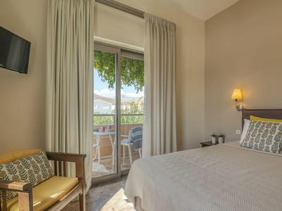 Chambre d'hôtel lumineuse avec lit double, fauteuils et balcon donnant sur des arbres verts.