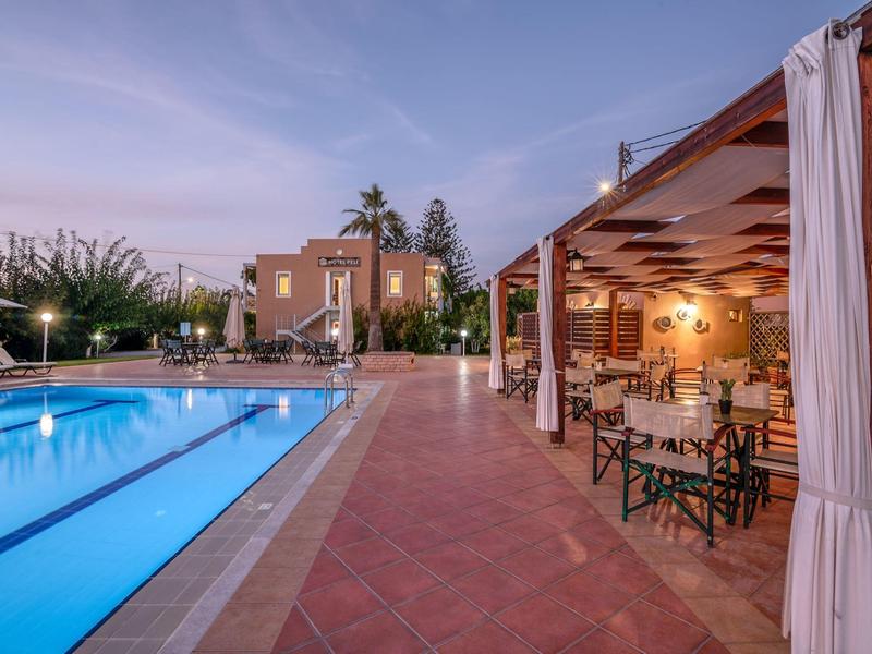 Vue en soirée d'une piscine éclairée à côté d'un restaurant extérieur couvert avec des tables et des chaises.