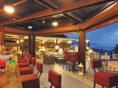 Offene Hotelbar mit roten Hockern und Sitzbereichen, beleuchtet in der Abenddämmerung am Meer.