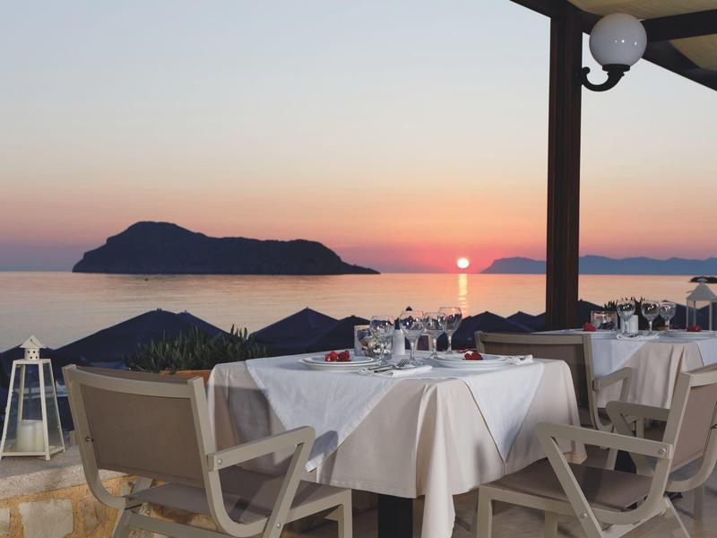Terraza de restaurante junto al mar con mesas preparadas al atardecer y vista a una isla.