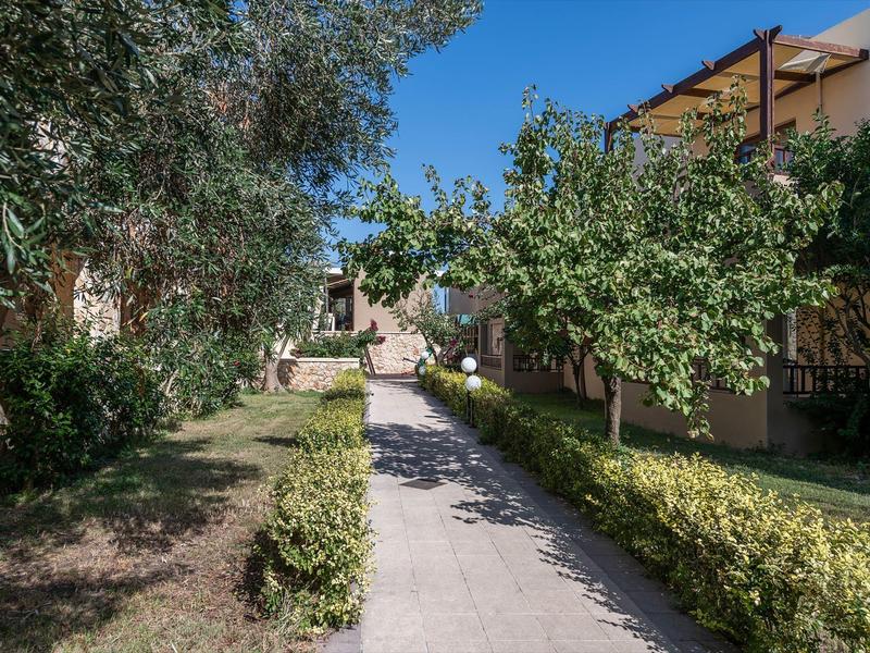 Camino entre setos verdes y árboles conduce a un edificio bajo un cielo azul.