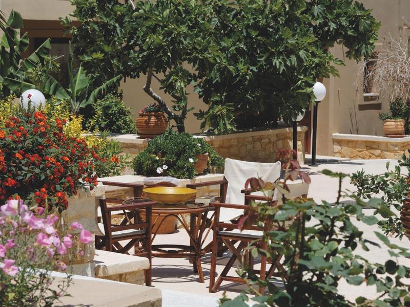 Terraza soleada con mesa y sillas de madera, rodeada de plantas y flores mediterráneas.