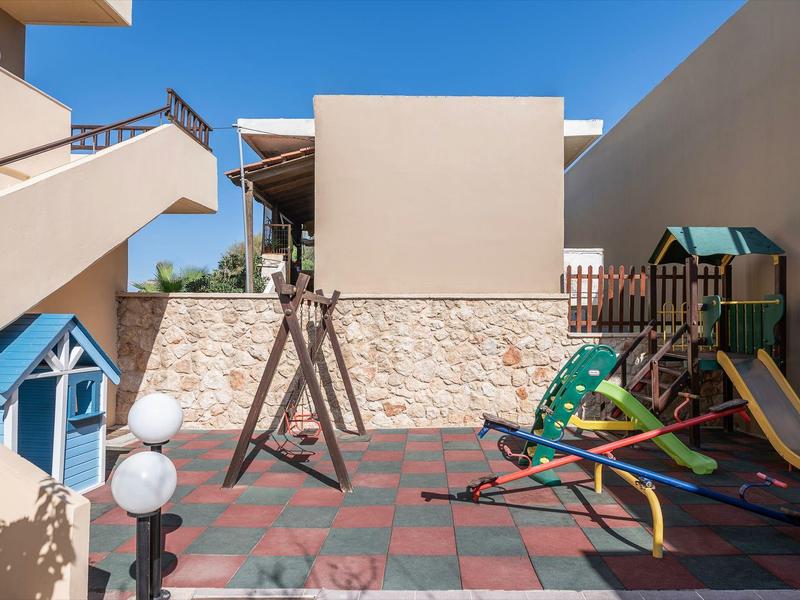 Un pequeño parque infantil soleado con tobogán, columpios y estructura para trepar en el patio del hotel.
