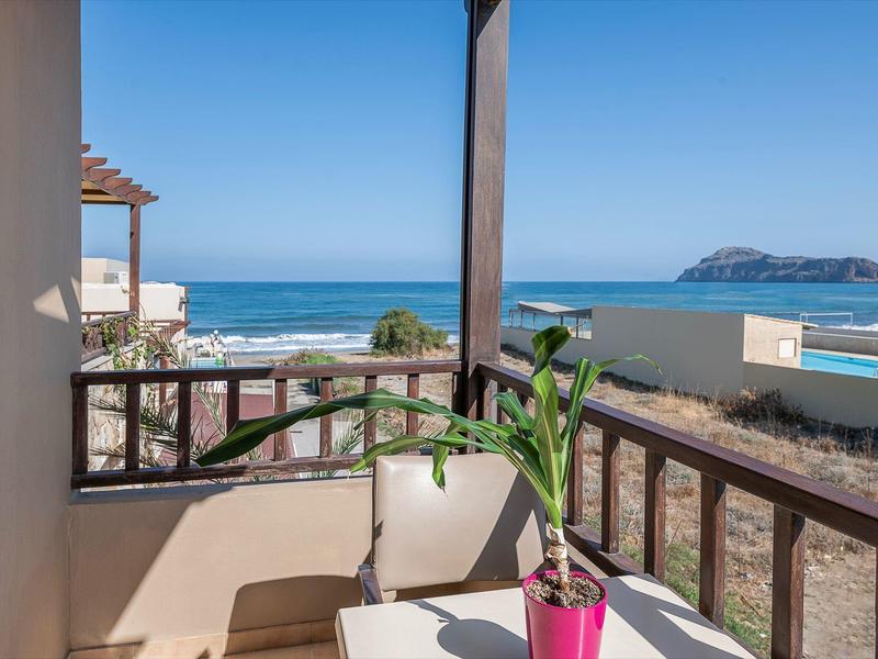 Balcón con sillas blancas y planta, vista a playa de arena y mar bajo cielo despejado.