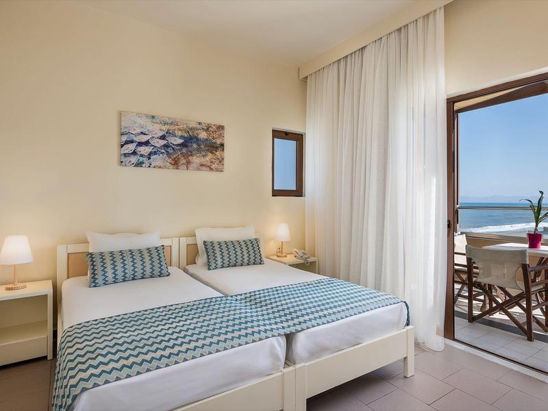 Habitación de hotel luminosa con dos camas, mesitas, lámparas y balcón con vista al mar.