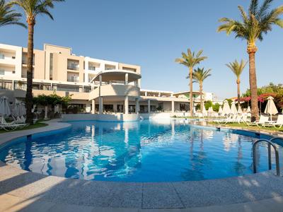 Runder Hotelpool mit Sonnenschirmen und Palmen, umgeben von Liegestühlen bei klarem Himmel.