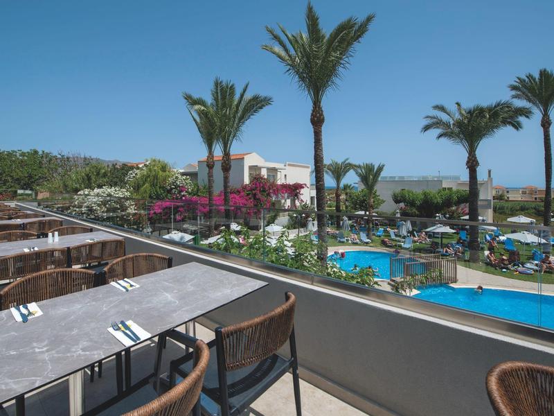 View from a terrace bar with tables overlooking a pool area with palm trees and garden