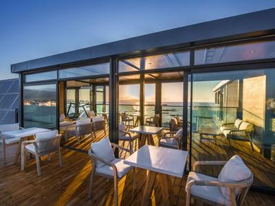 Terrasse mit Stühlen und Tischen vor einem modernen Gebäude mit Meerblick bei Sonnenuntergang.
