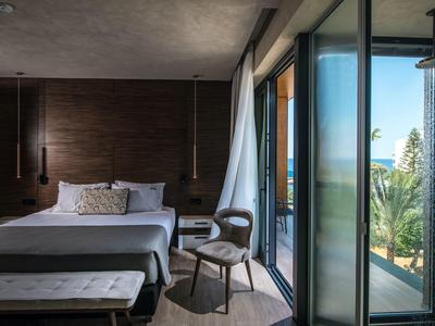 Modern hotel room with large bed, chair, and balcony access overlooking palm trees.