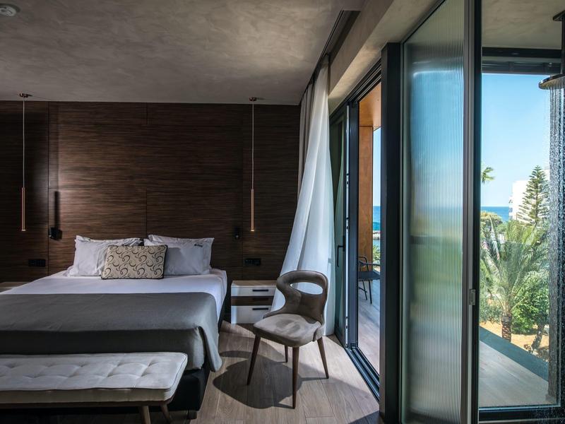 Modern hotel room with large bed, chair, and balcony access overlooking palm trees.