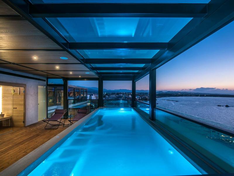 Modern hotel pool with glass roof and sea view at sunset.