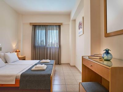 Jednoduše zařízený hotelový pokoj s manželskou postelí, psacím stolem a oknem s průhlednými závěsy.