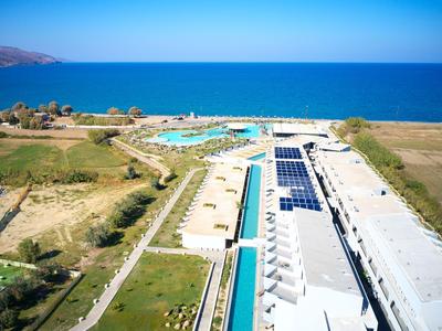 Luftaufnahme eines Hotels am Meer mit Pool und umliegender grüner Landschaft unter klarem Himmel.