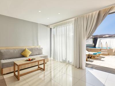 Helles Hotelzimmer mit grauem Sofa, Holztisch und Zugang zu Terrasse mit Pool und Sonnenliegen.