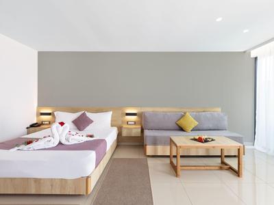 Helles Hotelzimmer mit Doppelbett, grauem Sofa, Holzmöbeln und großen Fenstern mit weißem Vorhang.