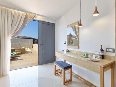 Heller Hotelraum mit Balkontür, Vorhängen, Tisch, Hocker und Blick auf Terrasse mit Meerblick.