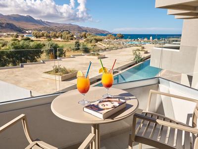 Balkontisch mit zwei Cocktails und Buch, Blick auf Meer, Strand und Berge bei sonnigem Wetter.