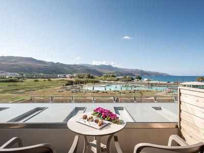 Blick von einem Balkon mit Tisch und Blumen auf eine grüne Landschaft, Pool und Berge im Hintergrund.