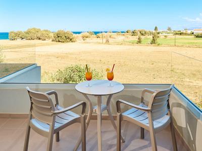 Balkon mit Tisch und zwei Stühlen, zwei Getränken, Blick auf eine weite Wiese und das Meer im Hintergrund.