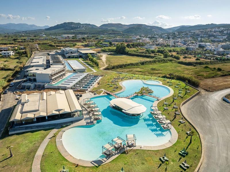 Groot, gebogen hotelzwembad met zonneterras en ligstoelen omgeven door groen landschap.