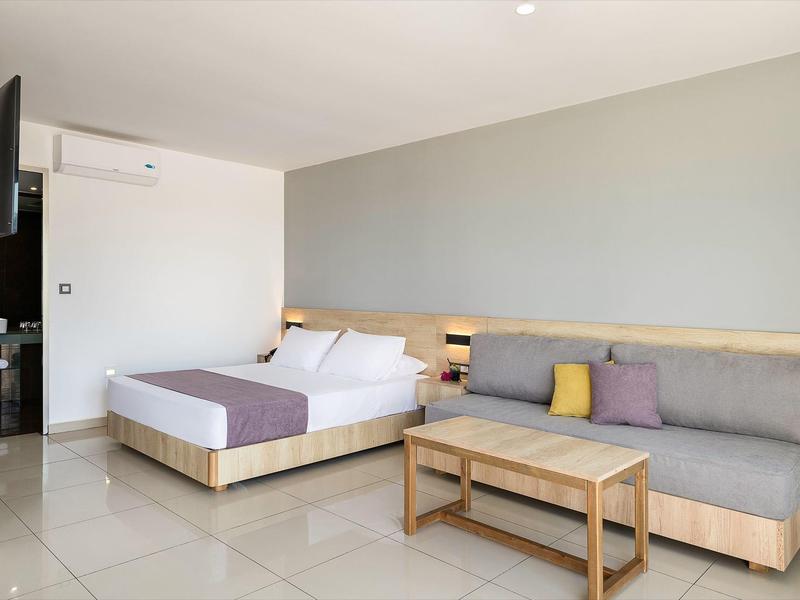 Modern hotelkamer met tweepersoonsbed, bank en kleine houten tafel in lichte omgeving.