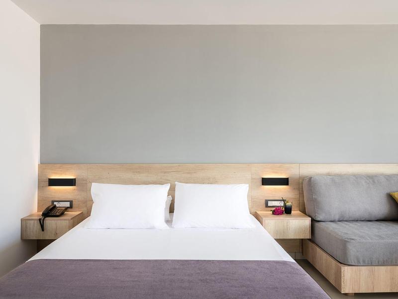 Modern hotelkamer met tweepersoonsbed, twee witte kussens, houten hoofdeinde, nachtkastjes en grijze bank.