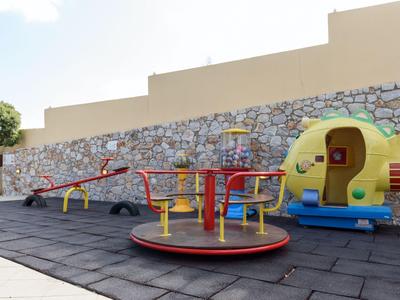 Spielplatz mit Karussell, Wippe und Spielhaus neben einer gepflasterten Mauer im Hotelbereich.
