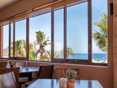 Blick durch große Fenster auf Palmen und Meer von einem Hotelrestaurant bei Tageslicht.