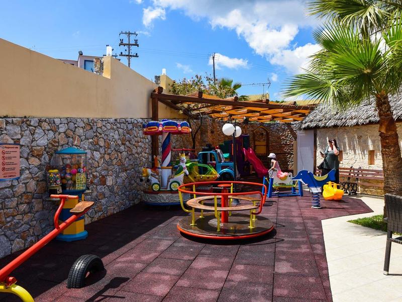 Children's playground with carousel, slide, and seating, surrounded by stone walls and palm trees.