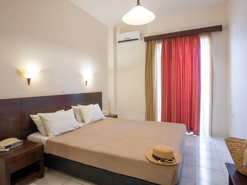 Bright hotel room with double bed, nightstands, air conditioning, and red curtains on the window.